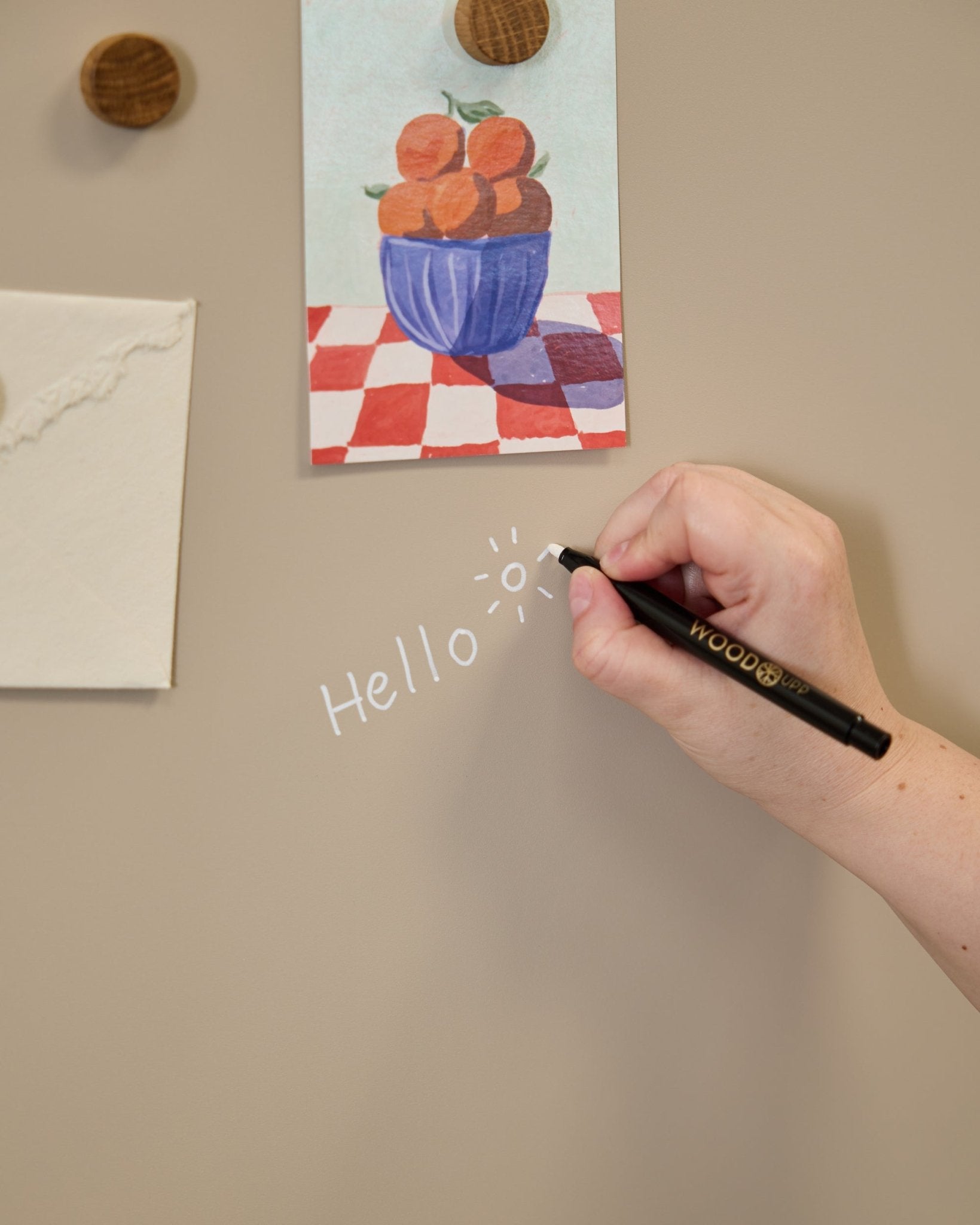 Une main écrit hello avec un dessin de soleil sur le Blackboard WoodUpp Norr, placé à côté d’illustrations fixées avec des aimants ronds sur un Panneau acoustique.